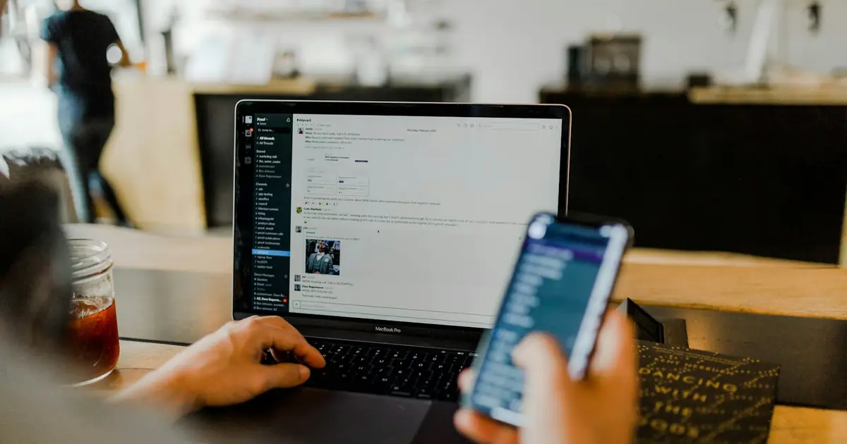 A person working at a laptop while holding a smartphone.